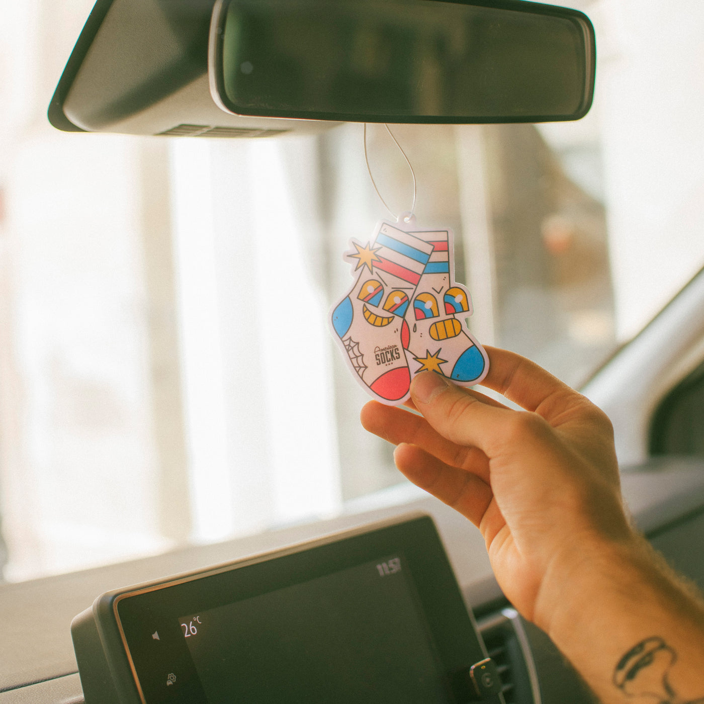 Hand holding a colorful air freshener hanging from a car's rearview mirror.