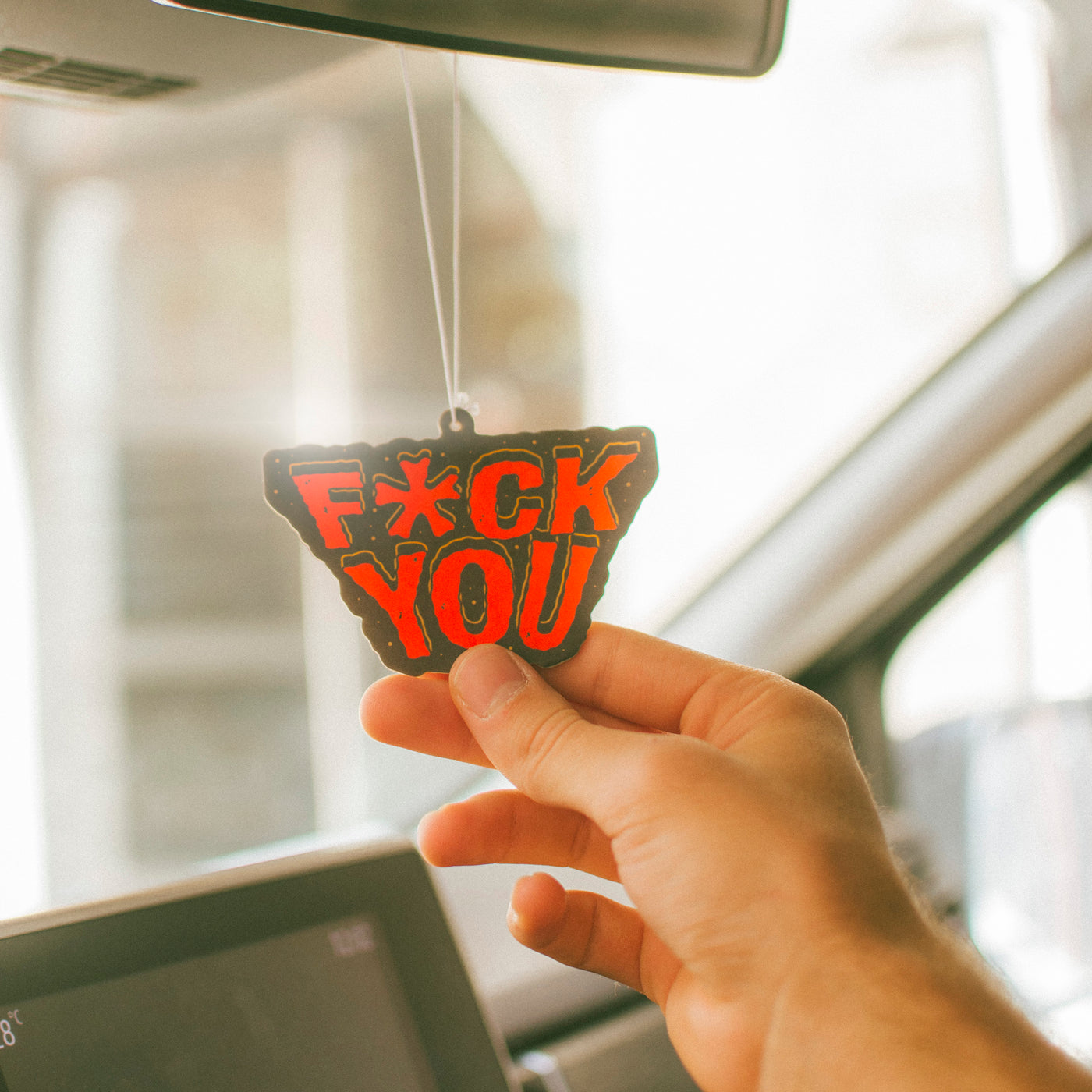 Hand holding a car air freshener with 'F*CK YOU' text inside a vehicle.